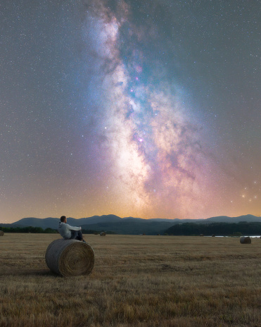 Yound adult sitting on a grass bale watching the night sky, milky way core visible