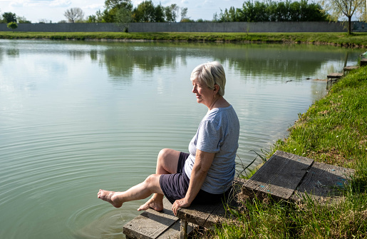 An elderly woman sits by the water with bare feet