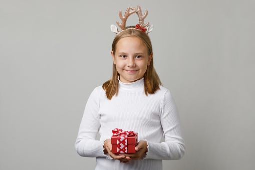 Beautiful child wearing white longsleeve t-shirt and Christmas hairband holding Xmas gift. Young cute girl Christmas portrait