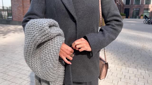 Close up of woman's hands fastening button on gray jacket, bundling up and holding warm knitted woolen scarf