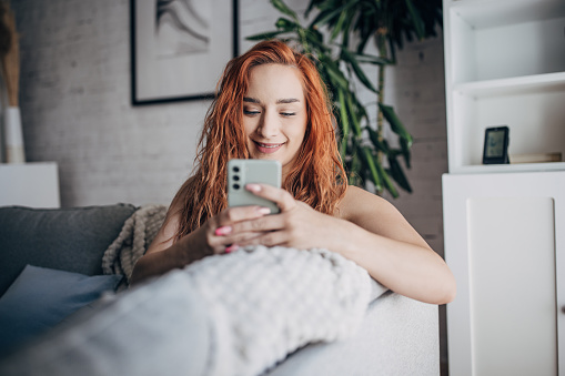 One woman, beautiful redhead woman using smart phone while sitting on sofa at home.