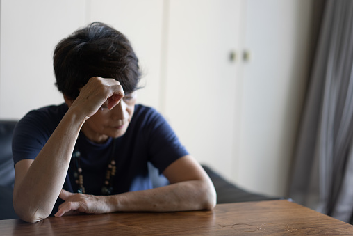 An elderly woman sits on a sofa and is distressed.
She is resting one elbow on the table.
Depression.
In the living room at home.
She is a Japanese woman in her seventies.
