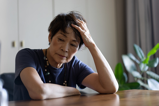 An elderly woman sits on a sofa and is distressed.\nShe is resting one elbow on the table.\nShe rakes up her hair with a melancholy expression.\nA sense of weariness. Lack of motivation.\nIn the living room at home.\nShe is a Japanese woman in her seventies.