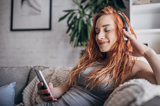 One woman, redhead woman using smart phone and listening music on headphones while sitting at home.