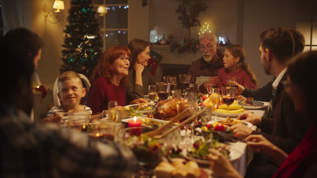 Grandfather Telling Funny Childhood Stories to His Young Granddaughter, Who is Sitting on a Lap at a Christmas Table with Diverse Family and Multicultural Friends. Festive Dinner Party in the Evening