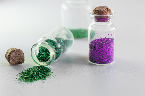 Glitter sales ban in Europe concept. Environmental protection. Purple and green glitter in glass bottles. Isolated on white background.