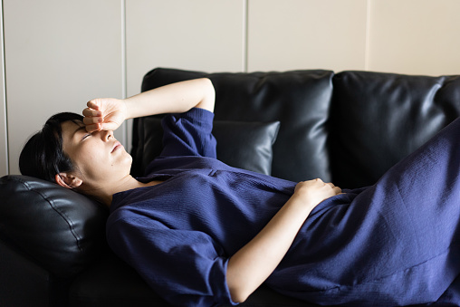 A woman lying down on a sofa.
A woman putting one hand on her forehead.
A woman holding her stomach with one hand.
A woman in the early stage of pregnancy.
A woman sweating cold sweat.