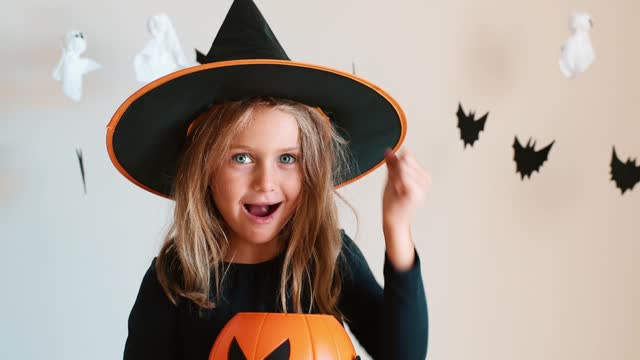 Video portrait happy excited face girl trick or treat Halloween surprise kid wearing dress witch hat costume party Halloween. Toddler holding pumpkin basket candies trick or treating