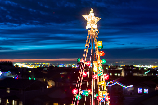 Close up of the top of an illuminated Christmas Tree decoration with a star and ornaments, with a community of homes and the Spokane Washington city skyline behind at twilight evening.