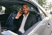 Smiling businessman talking phone with client while sitting on car backseats