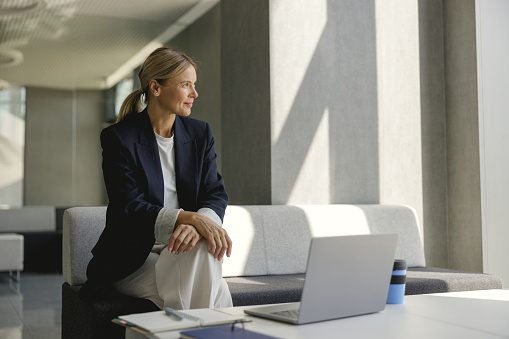 Smiling business woman working on laptop sitting on background of modern office and looks away