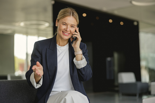 Cheerful businesswoman talking phone sitting on modern office background and looks away