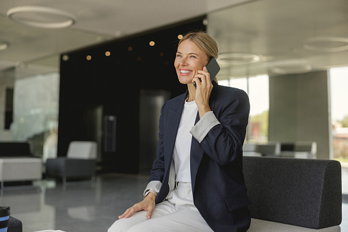 Smiling female entrepreneur talking phone sitting on modern office background and looks away