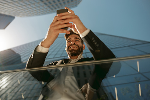 Bottom view of handsome entrepreneur using mobile phone standing on background of skyscrapers