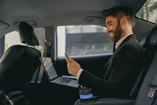 Handsome businessman in suit using personal computer and using mobile phone in taxi