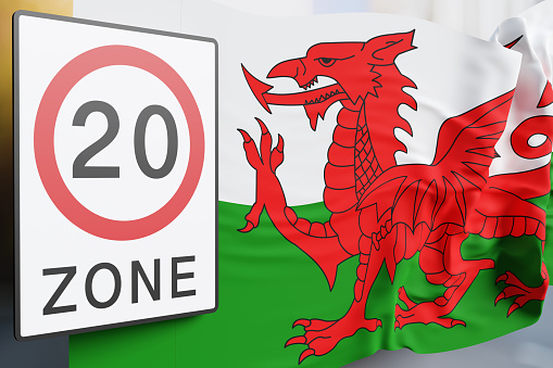 Road sign of 20mph zone and Welsh national flag in front of a street view. Illustration of the concept that Wales has lowered the speed limit on one-way roads in towns and cities