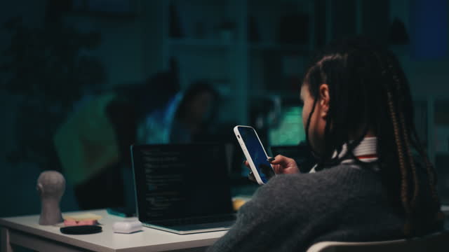 African American programmer scrolling phone while working late night in office