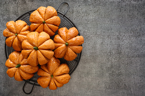 Stock photo showing close-up, elevated view of freshly baked, homemade, Halloween pumpkin designed rolls with green chilli pepper stalks on a circular, black cooling rack.