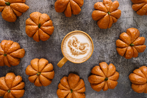 Stock photo showing close-up, elevated view of autumnal, Halloween scene of orange, coffee mug surrounded by homemade Halloween pumpkin designed bread buns glazed with apricot jam arranged on mottled grey background. The cup contains a hot Latte decorated with a scary ghoul face design milk foam art.