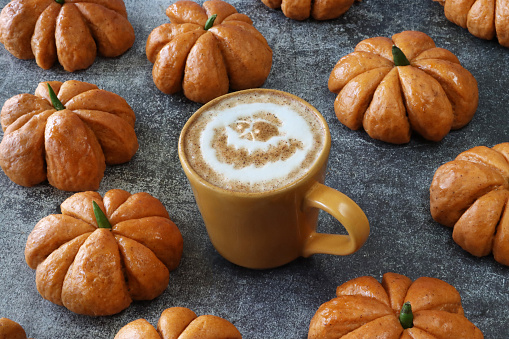 Stock photo showing close-up, elevated view of autumnal, Halloween scene of orange, coffee mug surrounded by homemade Halloween pumpkin designed bread buns glazed with apricot jam arranged on mottled grey background. The cup contains a hot Latte decorated with a ghost face design milk foam art.