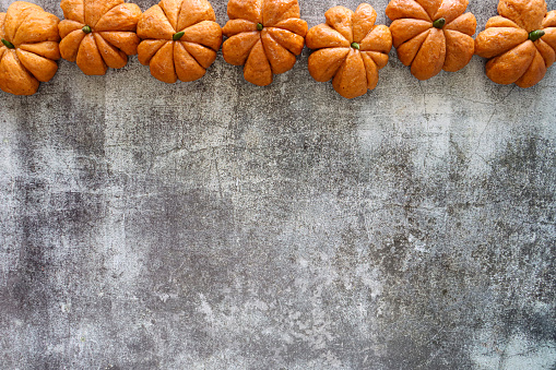 Stock photo showing close-up, elevated view of border of freshly baked, homemade bread pumpkin designed rolls with green chilli pepper stalks made into a picture frame, against a grey background.