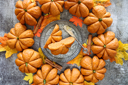 Stock photo showing close-up, elevated view of Halloween wreath of freshly baked, homemade bread pumpkin designed rolls with green chilli pepper stalks against a grey background.