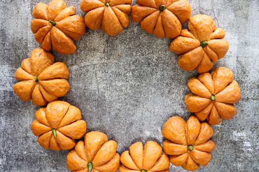 Stock photo showing close-up, elevated view of Halloween wreath of freshly baked, homemade bread pumpkin designed rolls with green chilli pepper stalks against a grey background.