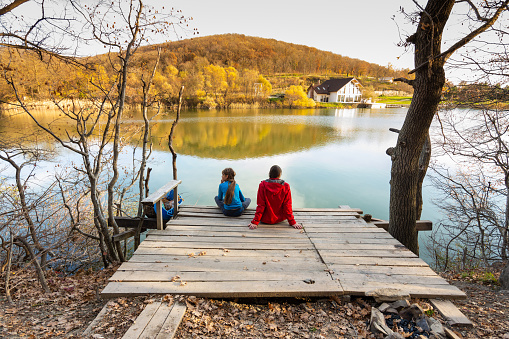 A girl and a girl are sitting on a wooden bridge near a mountain lake, a view from the back of people