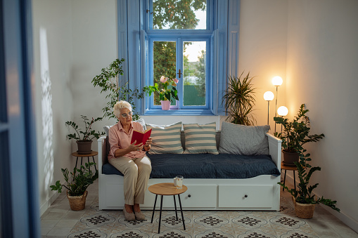 Caucasian elderly woman reading her book and sitting on the sofa of her living room while being relaxed and cozy