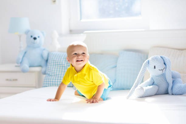 Baby playing on parents bed. Cute funny little boy learning to crawl in white sunny nursery. Infant and toddler room interior. Bedding and toys for kids. Children play at home. Baby bedroom.