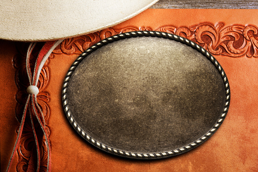 A large belt buckle and a leather stampede string of a cowboy hat rest on top of a tooled leather surface.