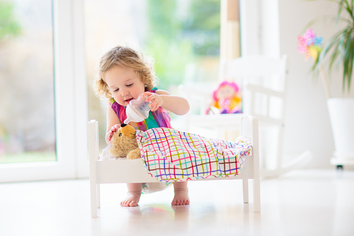Child playing with teddy bear. Kids play. Little girl feeding doll with milk bottle. Stuffed animal in toy bed. Role game for young kids. Children play. Nursery interior.