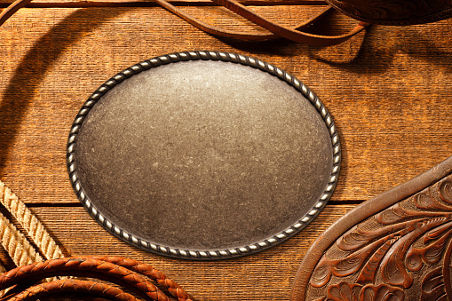 A belt buckle framed by lariat,saddle flap, and leather riding tack on a weathered wood surface.