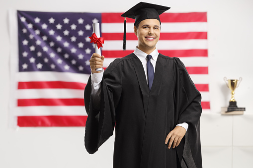 Young man in a graduation gown and cap holding a diploma in front of USA flag