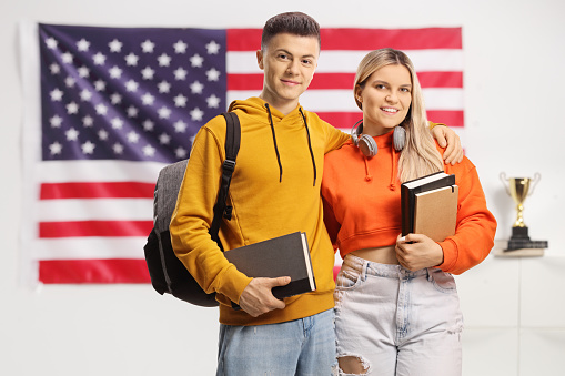 Students with backpacks and books standing in front of a USA flag