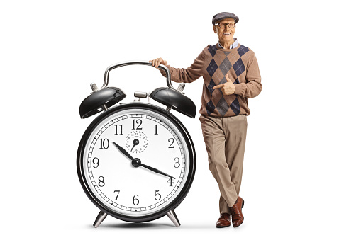 Elderly man leaning on a big alarm clock and pointing isolated on white background
