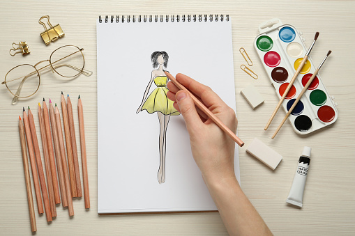 Fashion designer creating new look. Woman drawing sketch in pad with pencil at white wooden table, top view