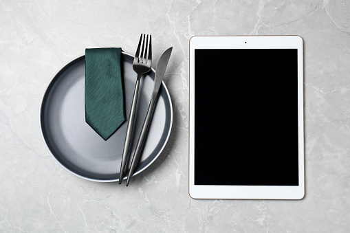 Business lunch concept. Plate, cutlery, tie and tablet with space for text on light gray marble table, flat lay