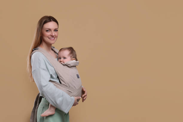 istock Mother holding her child in sling (baby carrier) on light brown background. Space for text 1707074195