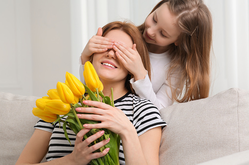 Daughter covering mother's eyes with her palms and congratulating with bouquet of yellow tulips at home