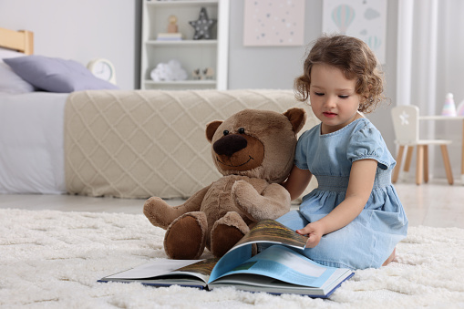Cute little girl with teddy bear and book on floor at home