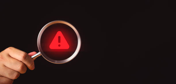 istock Hand holding magnifier glass with red triangle caution warning sign on isolated black background with copy space. Cyber criminals focus on the notification error and maintenance concept. 1707139863
