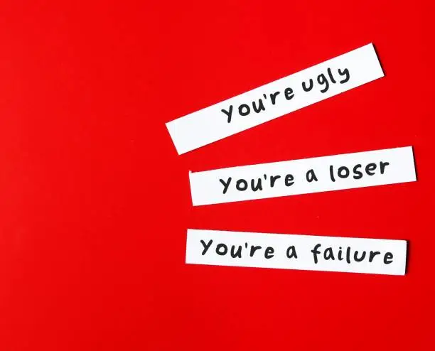 On copy space red background, paper pieces written You're ugly , You're a looser, You're a failure - verbal abuse concept - negative demeaning words abuser uses to gain or maintain power and control over victims