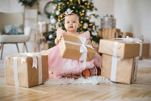 Front view of caucasian charming girl sitting on wooden sleigh and holding craft wrapped gift in hands. Attractive little baby in pink dress playing with presents on background of Christmas tree.