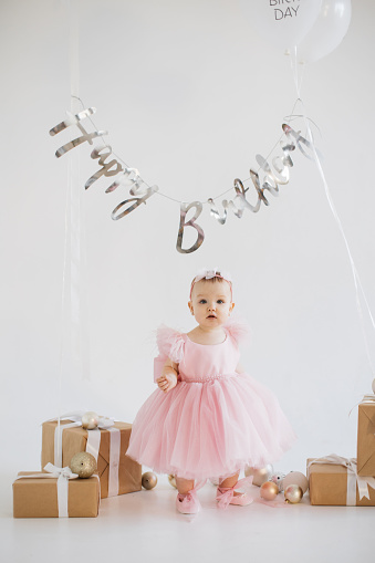 Playful little girl wearing finest party dress standing alone in studio adorned with festive decorations. Charming caucasian baby girl with sincere emotions celebrating birthday party.
