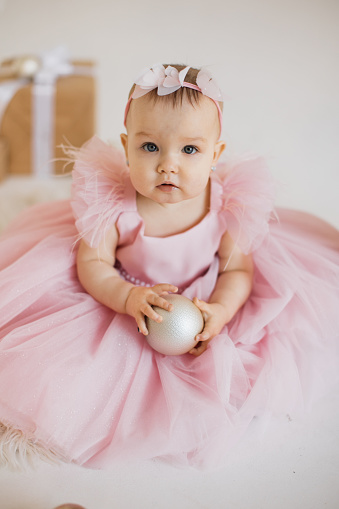 Small beautiful princess dressed in festive wear sitting with white ball in hands in decorated studio. Attractive pretty toddler with cute headband looking aside and enjoying birthday party.