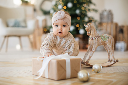 Attractive caucasian baby girl reaching for craft gift in studio decorated with garlanded Christmas tree. Charming little girl preparing to celebrating first New Year in life.