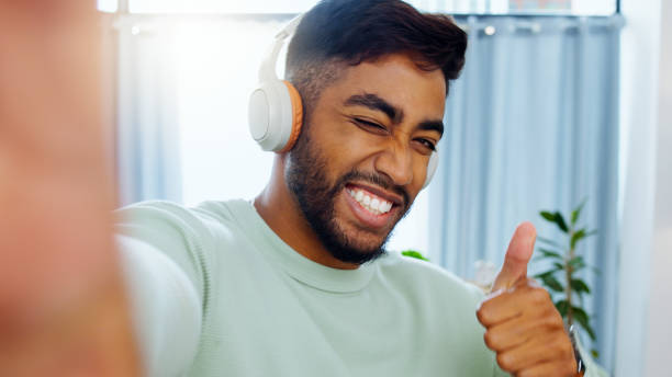Selfie, man and smiling thumbs up emoji headphones for music playlist streaming and cheerful mood in home. Wellness peace sign, audio and happy male ready to dance photograph with smile wink Selfie, man and smiling thumbs up emoji headphones for music playlist streaming and cheerful mood in home. Wellness peace sign, audio and happy male ready to dance photograph with smile wink young man wink stock pictures, royalty-free photos & images