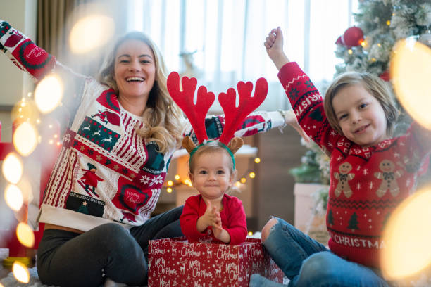 mutter und ihre kinder mit erhobenen armen blicken am weihnachtstag zu hause in die kamera - sibling baby three people baby girls stock-fotos und bilder