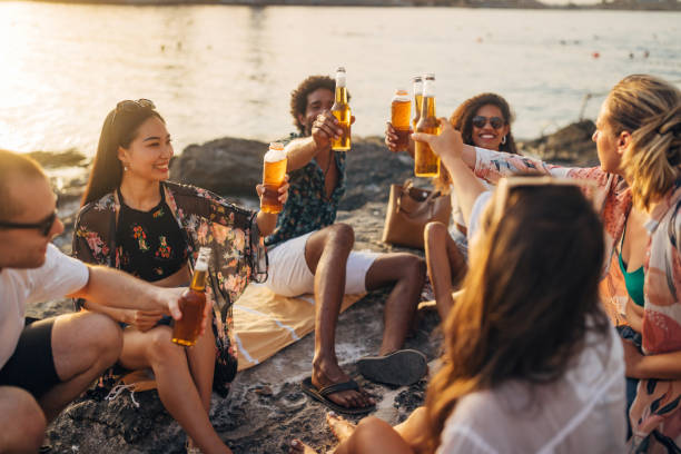 Nature, cheers and diversity, friends in sunset in celebration and medium group of people on happy summer holiday together Diverse group of friends sitting on the beach, drinking beer and toasting friendship and a joint, group vacation. Beer, men and women relax and toast the vacation golden hour drink stock pictures, royalty-free photos & images